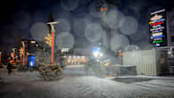 A snowplough at work on a city square surrounded by snow-covered Christmas trees and shop signs.