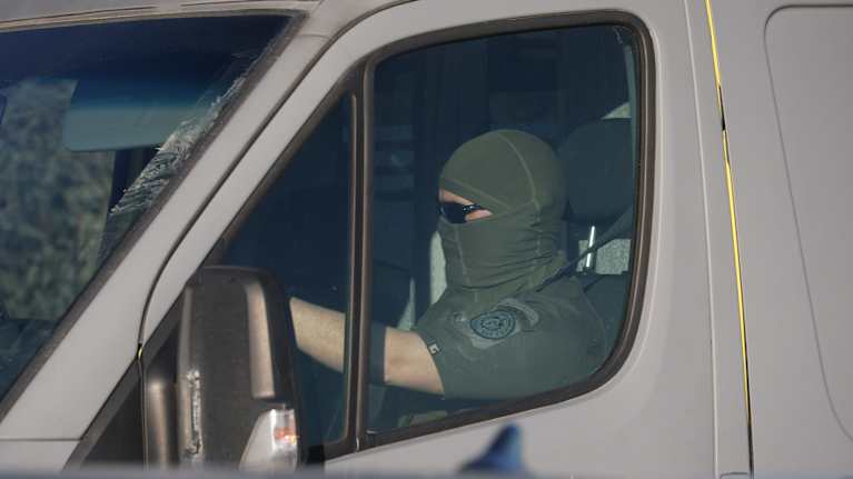 A man driving a grey van, wearing short-sleeved green uniform and balaclava.