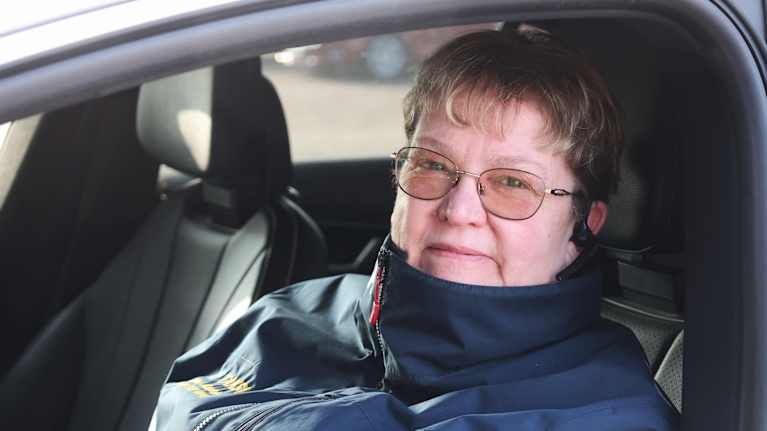 En person sitter i en bil, klädd i en mörk jacka med ett taxiemblem på vänster sida.