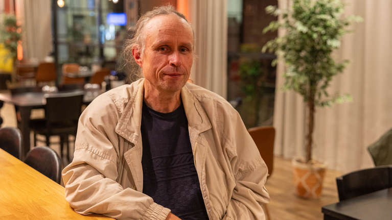 En man med ljust hår sitter vid ett träbord i en restaurangmiljö med stolar, bord och växter i bakgrunden.