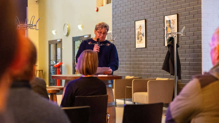 En person står vid ett bord och talar i mikrofon inför sittande publik i ett rum med grå tegelvägg.