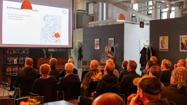 En person håller en presentation inför en sittande publik; en karta och texten "Esiintymisalueella" syns på en projicerad skärm.