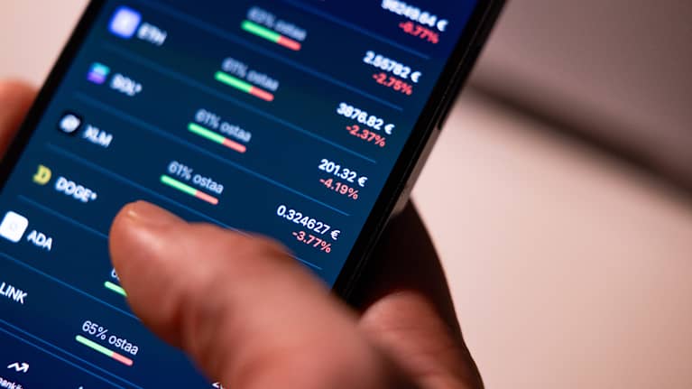 Close up photo of a hand holding a smartphone which is displaying cryptocurrency prices.