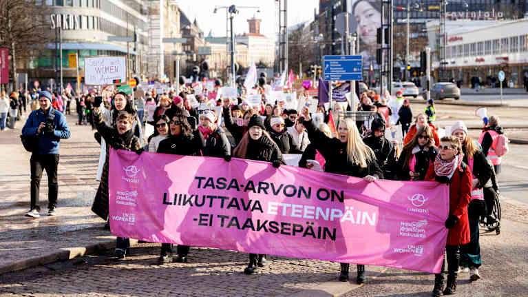 Suuri joukko ihmisiä marssii kadulla pidellen plakaatteja.