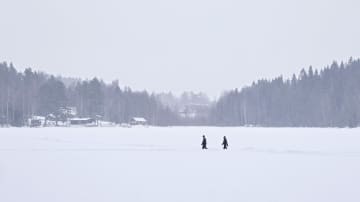 Lunta sataa. Maisemakuva järvelle, jossa kaksi  ihmistä kävelle. Taustalla rakennuksia. 