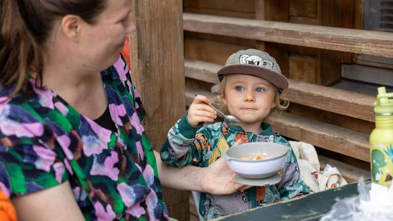 En kvinna hjälper ett barn att äta från en skål vid ett bord utomhus. Barnet har en keps på sig och håller en sked.