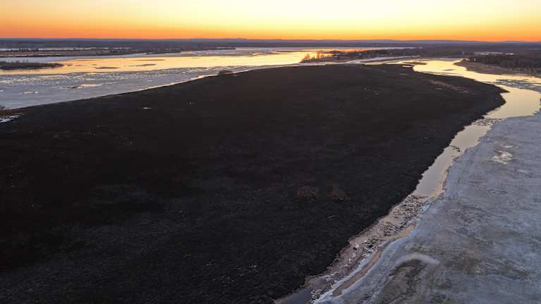 An aerial photo of a flat, charred island after a wildfire. The evening sky is glowing orange and yellow.