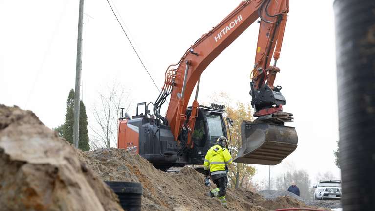 En person i varselkläder står i en grop bredvid en grävmaskin med texten "HITACHI" på armen.