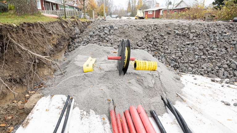 En byggarbetsplats med uppgrävd väg, grus, markrör och ledningar i rött, svart och gult samt hus i bakgrunden.