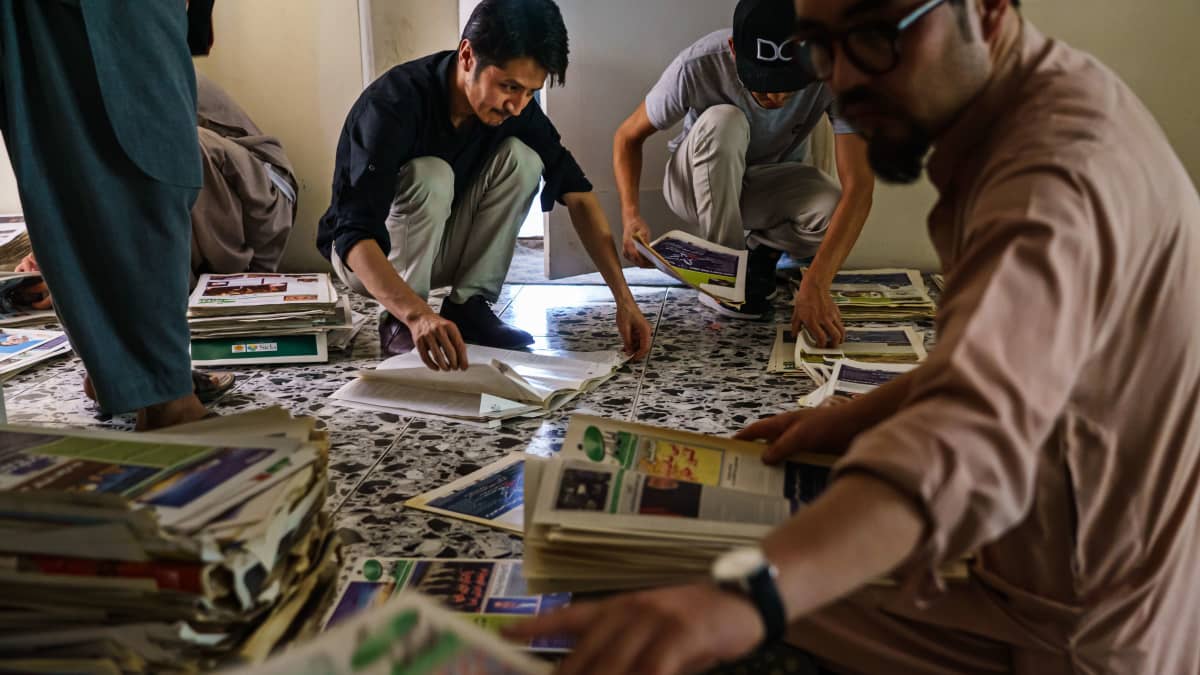 Länsimaisiin vaatteisiin pukeutuneet miehet lajittelevat sanomalehtipinoja lattialla Kabulissa Afganistanissa.