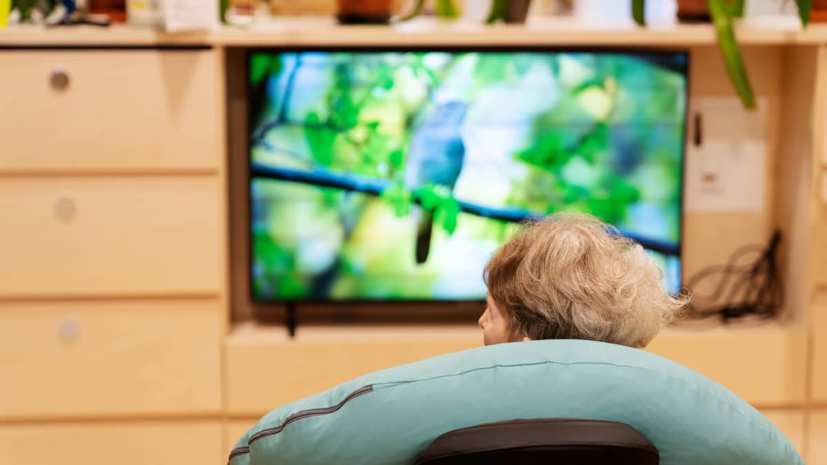 Vanha ihminen istuu katsomassa televisiota, hän on selin kameraan.