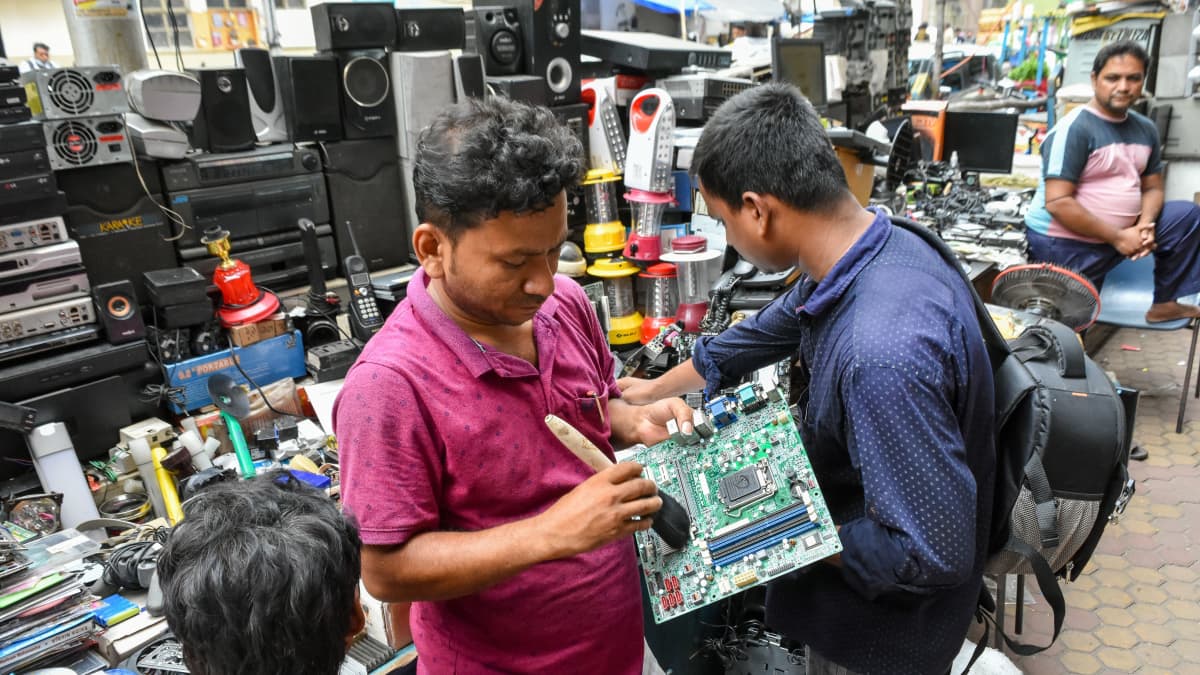 Punapaitainen intialainen mies puhdistaa tietokoneen emolevyä harjalla, taustalla erilasita elektroniikkaromua. 