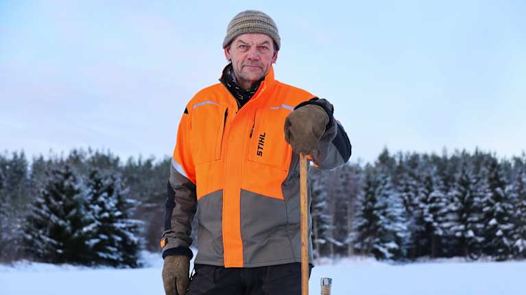 En man iklädd en orange arbetsjacka står utomhus på en snöig åkermark.