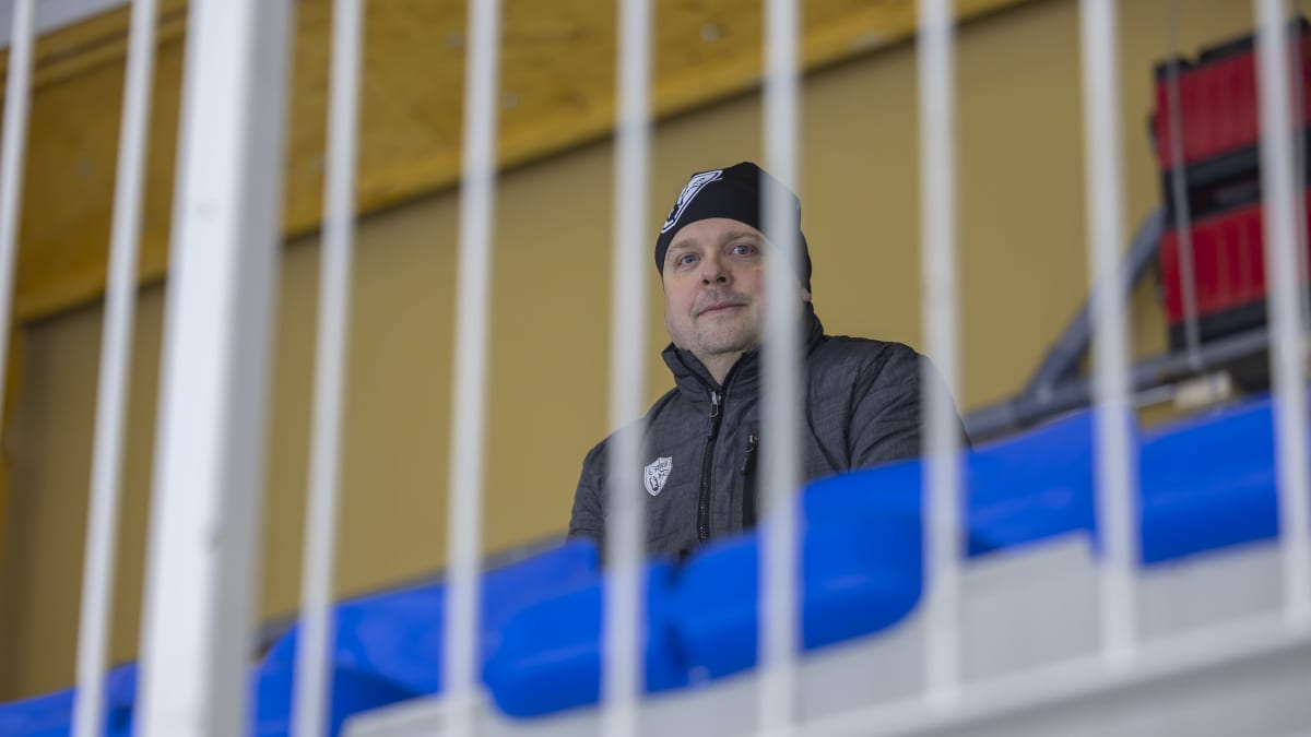 Janne Saarela istuu jäähallin katsomossa ja katsoo suoraan kameraan. Etualalla kaiteita epätarkkana.