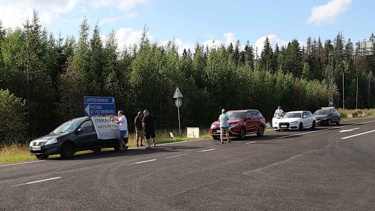 Ihmisiä seisomassa tien vieressä itaraja auki -banderollilla koristellun auton edessä.