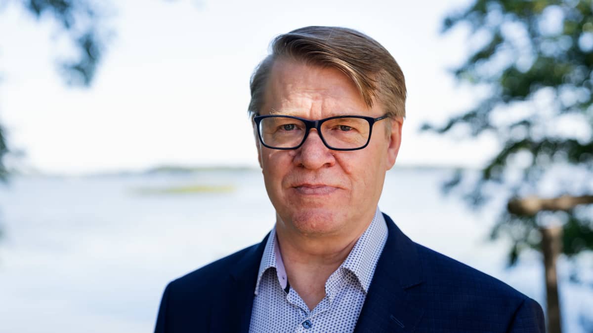 Silmälasipäinen mies katsoo suoraan kameraan, taustalla järvimaisemaa.