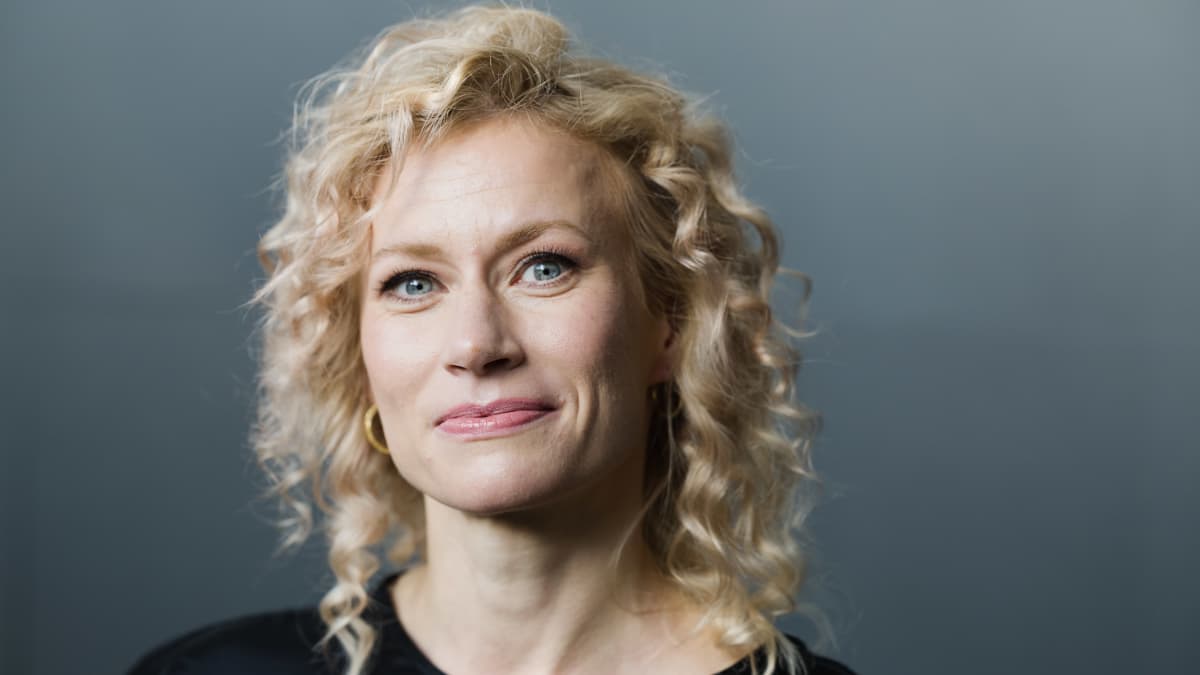 Minka Kuustosen potretti. Katsoo suoraan kohti kameraa, harmaa neutraali tausta.