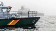A green coast guard boat at sea in foggy conditions, with a large oil tanker seen in the background.