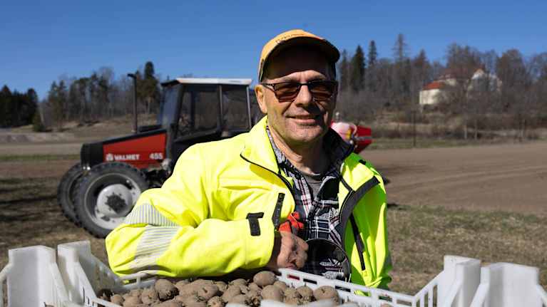 Jordbrukaren, potatisodlaren Claes Lindberg på en åker, lutad mot lådor med sättpotatis. En traktor i bakgrunden. 