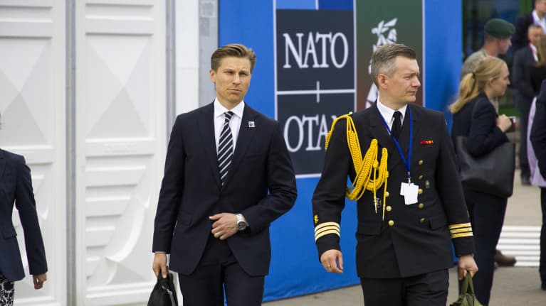 Antti Häkkänen, in a dark suit and striped tie and carrying a briefcase, walks with an aide in military uniform past a sign reading NATO / OTAN.