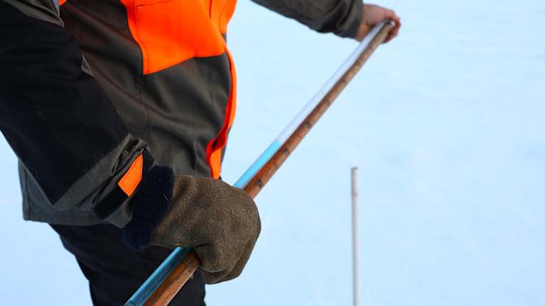 En man håller i en mätpinne och ett rör med en blå vätska i som frusit, som visar hur djup tjälen är. Bakgrunden är snötäckt mark.