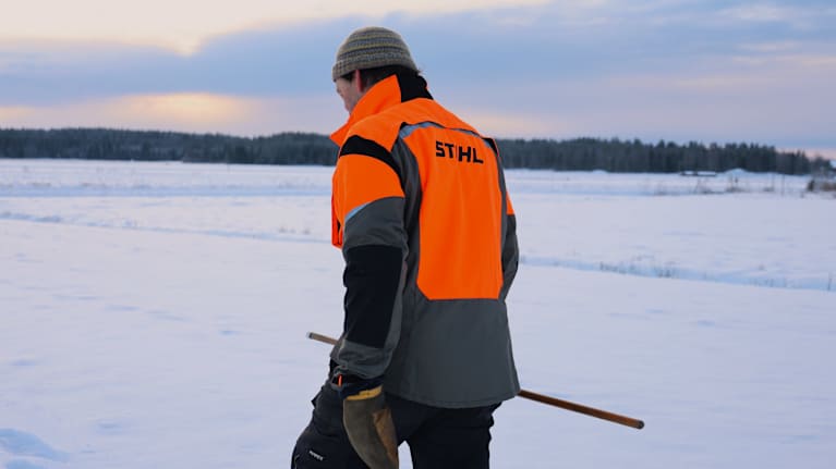 En man iklädd en orange reflexjacka går på en snöig åkermark.
