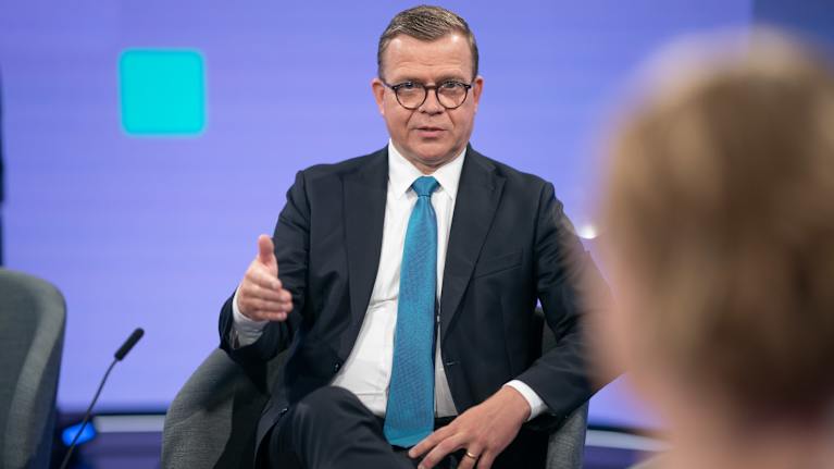 A man in a dark suit, blue tie and brown horn-rimmed glasses sat in a television studio and spoke with one hand half raised.