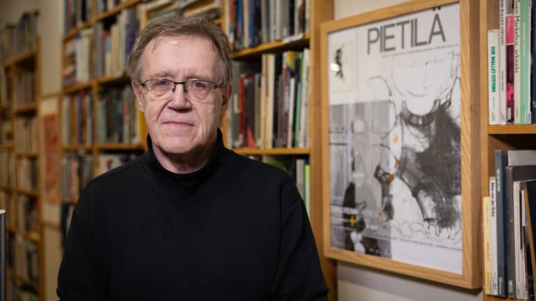 Photo of a grey-haired man wearing a dark shirt and wearing glasses looking into the camera, with bookshelves in the background.