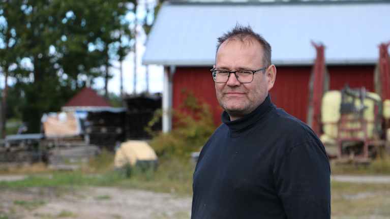 En man med kort hår och glasögon. I bakgrunden syns en röd lada.