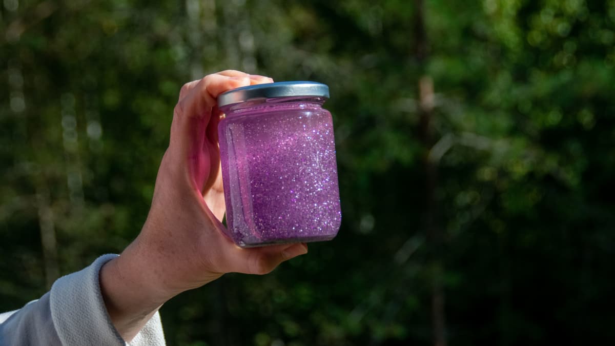 En hand håller i en glasburk fylld med rosa glitter och vatten.