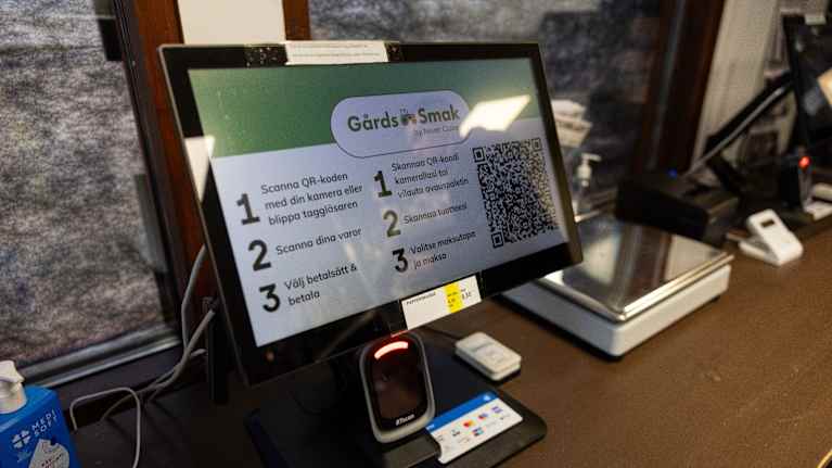 En datorskärm med instruktioner och en QR-kod står på en disk bredvid en betalterminal och en våg.