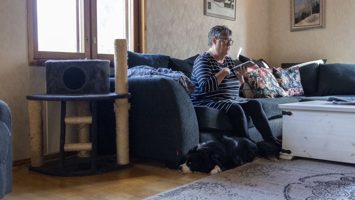 Kaija Hynninen istuu olohuoneensa sohvalla ja puhuu kaiuttimella olevaan puhelimeensa. Lattialla makaa mustavalkoinen koira. Sohvan vieressä on kissan raapimispuu