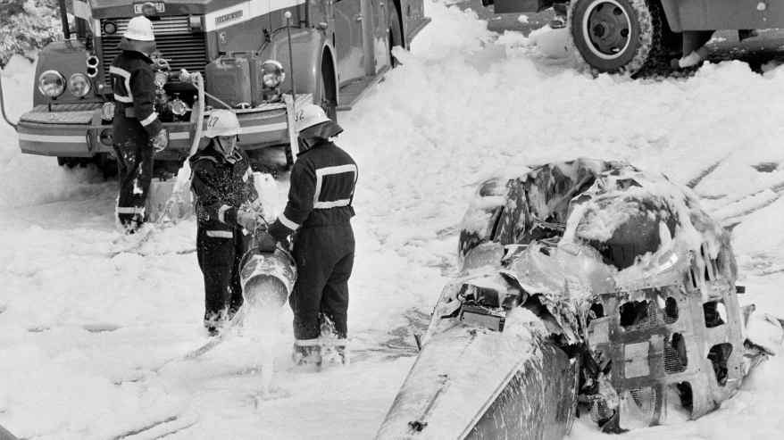 Pelastushenkilöitä lumessa pudonneen helikopterin vieressä. Mustavalkoinen kuva.