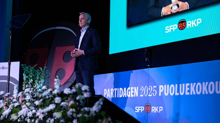 På bilden ser vi en man som står på en scen och talar vid ett evenemang. I bakgrunden finns skärmar med texten "PARTIDAGEN 2025 PUOLUEKOKOUS" och logotypen för SFP.