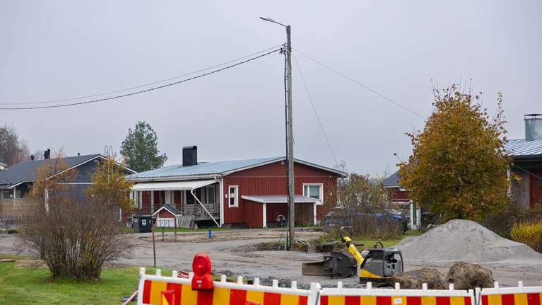 En grävmaskin och jordhögar syns bakom avspärrningar vid en väg intill röda bostadshus. Elledning.