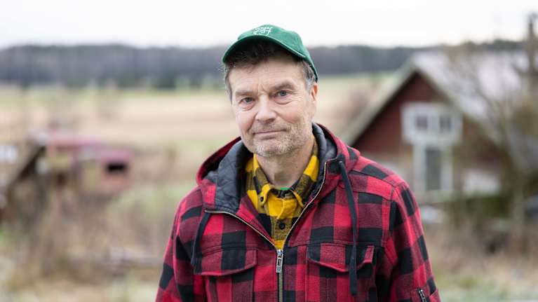 En person klädd i en röd och svart rutig jacka och en grön keps står utomhus på landsbygden med ett hus i bakgrunden.