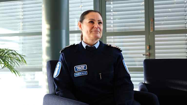 En polis i uniform. Hon sitter på en stol i ett kontorsutrymme. Uniformen har polisens symboler och epåletter på axlarna.