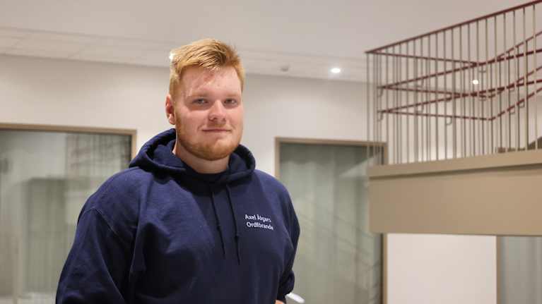 En person, Axel Ålgars, med rödblont hår står inomhus vid ett rött räcke.