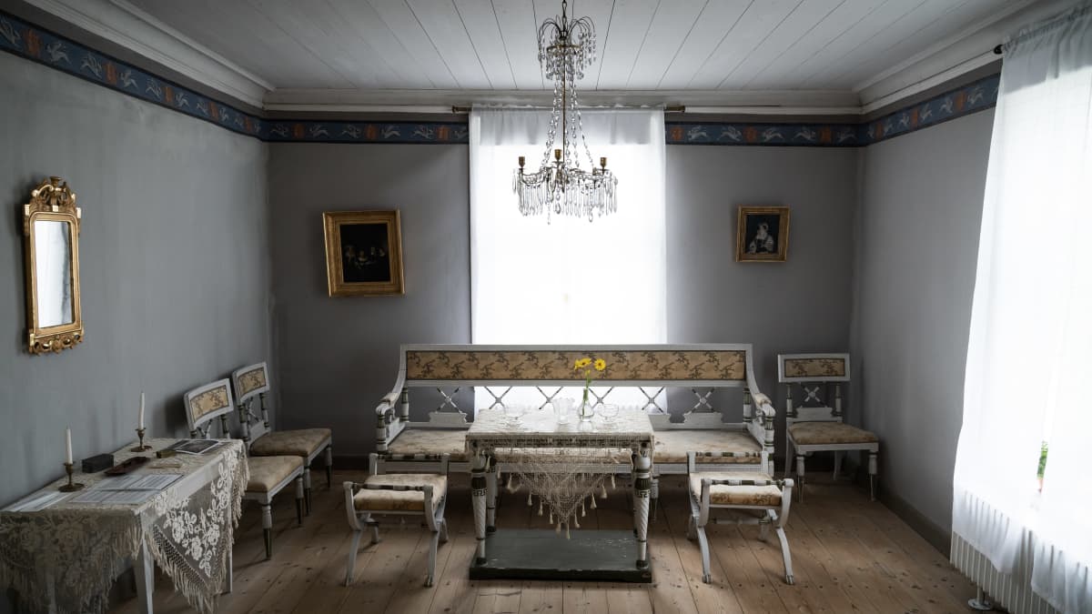 Sali Kuddnäsin kartanossa. Salissa on vanha kalusteryhmä, pöydillä on pitsiliinat, katossa on kattokruunu. Seinillä on kaksi taulua ja peili koristeellisissa kehyksissä. Kahdessa korkeassa ikkunassa on vaaleat valoverhot.