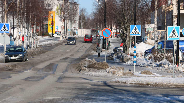 Stadscentra. Tre bilar kör på en väg och på trottoaren går några människor. Det är snö på marken. I förgrunden syns också ett flertal trafikmärken.