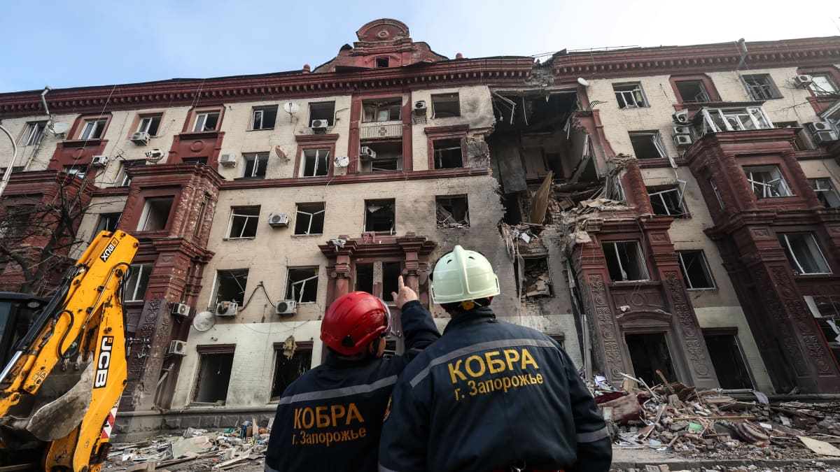 Kaksi kypäräpäistä ihmistä katsoo pahoja vaurioita saanutta rakennusta.