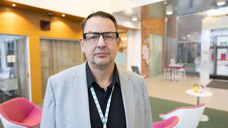 En man med glasögon och grå kavaj står i ett ljust kontorsutrymme med rosa och vita stolar.