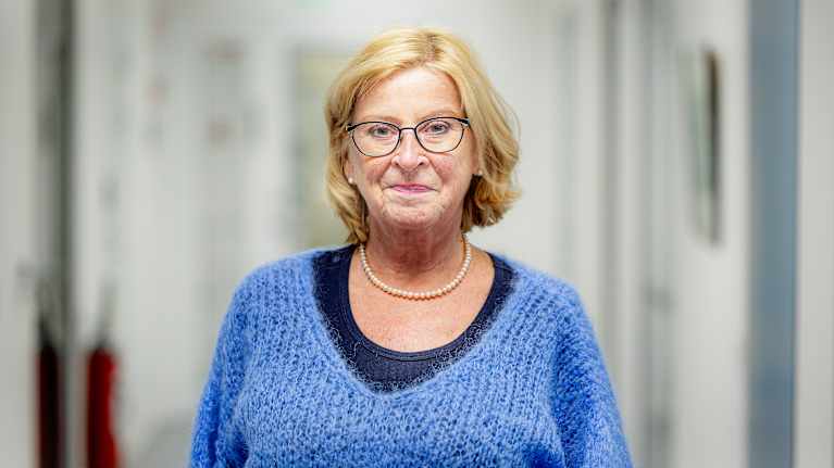 Kvinna i blå ylletröja står i en kontorsmiljö. Hon har ljust hår och glasögon.