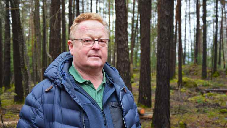 En man står i en skog iklädd en blå jacka och en grön skjorta.