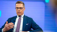 A man wearing glasses, a blue jacket and a purple tie sits in a TV studio, speaking with a serious expression and gesturing with one hand.