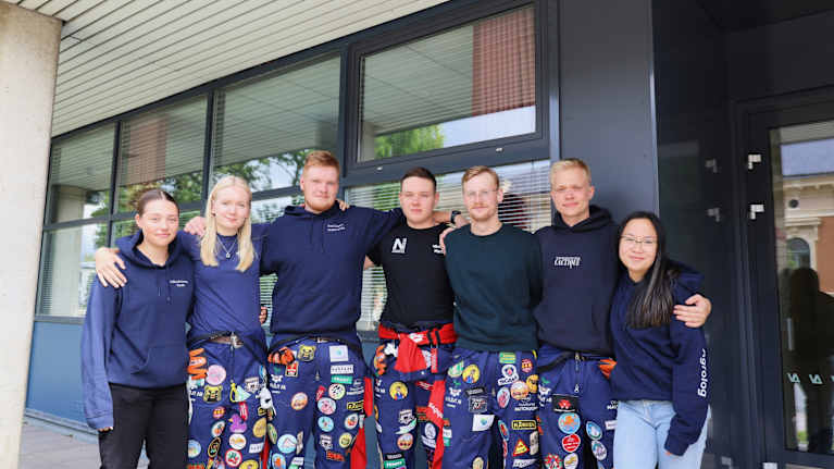 Sju personer står utomhus framför en byggnad, flera bär mörka huvtröjor och färgglada overaller med märken.