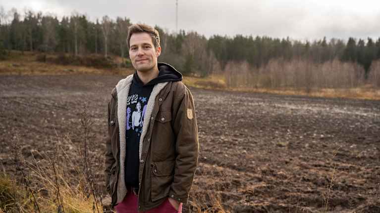 Man som ler mot kameran med plöjad brun åker i bakgrunden. 