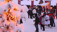 A snow-covered tree decorated with Christmas lights in the foreground, with many people wearing winter clothes in the background.