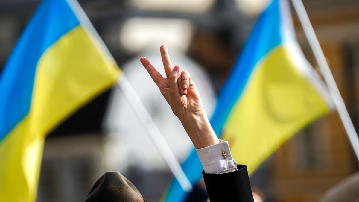 Photo shows a person making a peace sign symbol with two fingers, with two Ukrainian flags in the background.