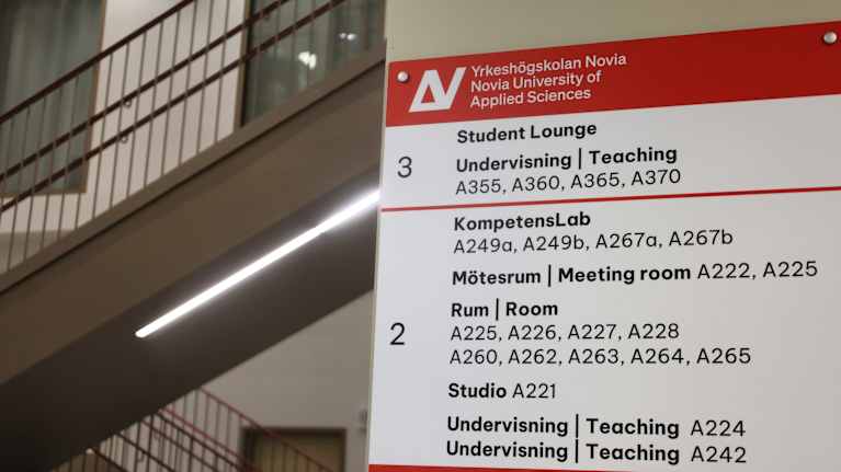 En vägvisningsskylt på Yrkeshögskolan Novia visar rumsnummer och funktioner på svenska och engelska.